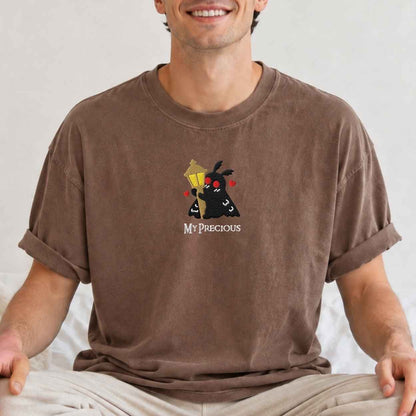 A man wearing a brown short-sleeve t-shirt with a graphic of a black moth holding a yellow street lamp and the text 'MY PRECIOUS' underneath, paired with beige pants against a light background.
