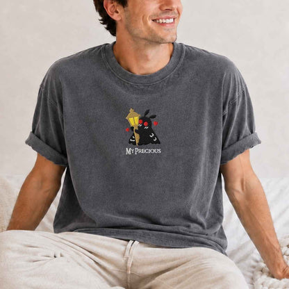 A man wearing a dark gray washed t-shirt with a cartoon graphic of a black moth holding a gold chain and red roses, with the text 'MY PRECIOUS' underneath. He is sitting on a bed with light-colored sheets.