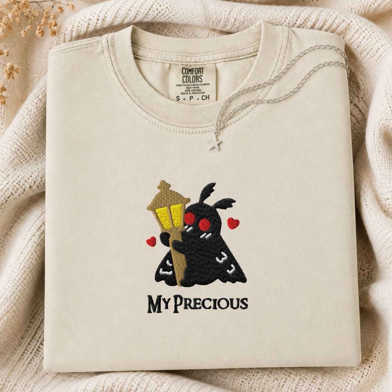 A folded cream-colored tshirt with a graphic of a black cartoon character holding a yellow street lamp and red hearts, with the text 'MY PRECIOUS' underneath. The tshirt is displayed on a cozy cream knit blanket with dried flowers in the corner.