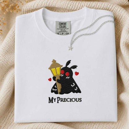 A folded slate white t-shirt with a custom embroidered design of a black moth holding a golden street lamp, surrounded by red hearts and the text 'MY PRECIOUS' in black. A silver star necklace is draped over the collar.