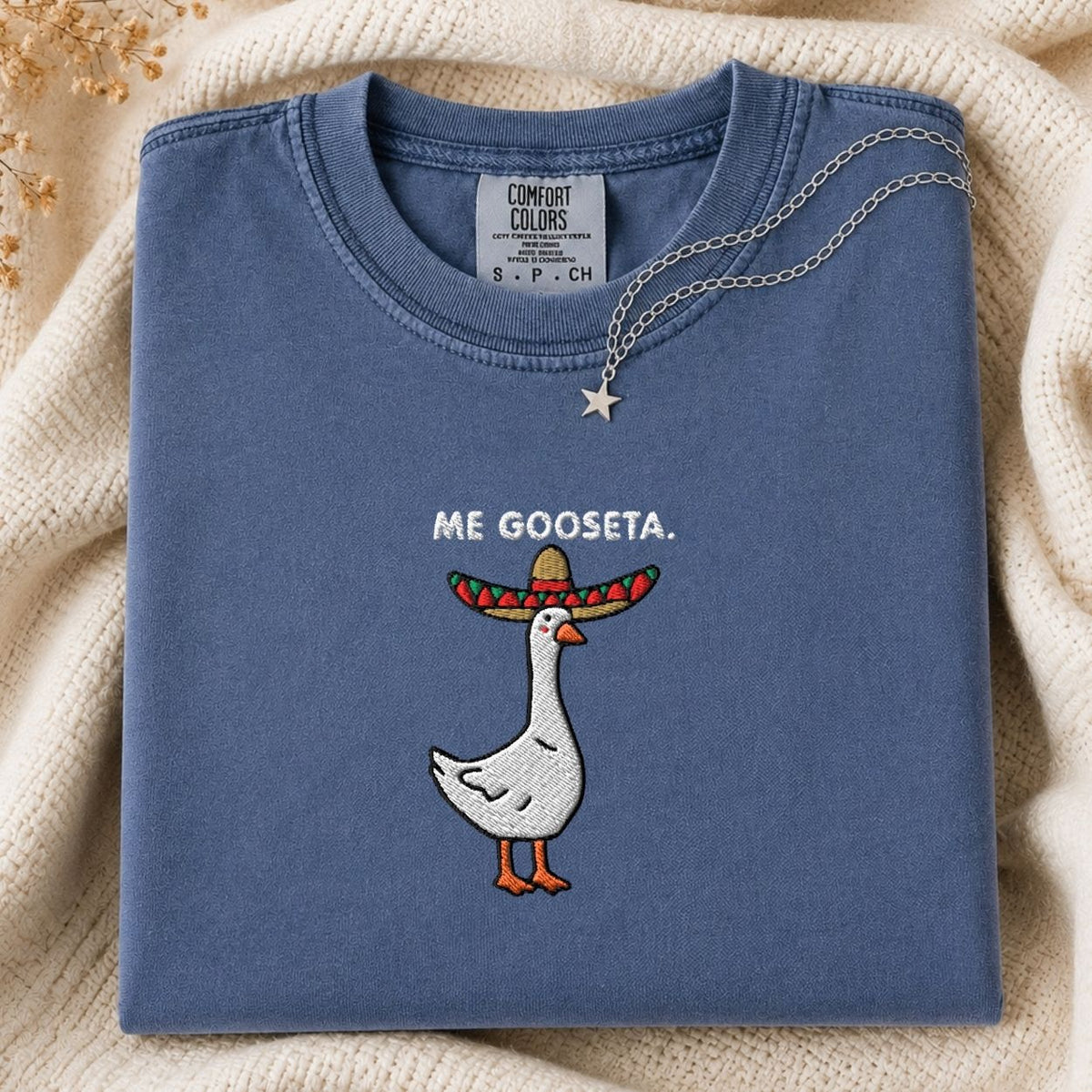 A folded slate blue t-shirt with a graphic of a white goose wearing a colorful sombrero and the text 'ME GOOSETA.' on the chest, styled on a cream knit blanket with a silver necklace.