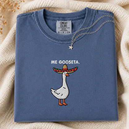 A folded slate blue t-shirt with a graphic of a white goose wearing a colorful sombrero and the text 'ME GOOSETA.' on the chest, styled on a cream knit blanket with a silver necklace.