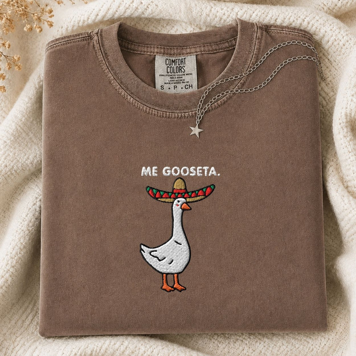 A folded brown Comfort Colors t-shirt with a graphic of a white goose wearing a colorful sombrero and the text 'ME GOOSETA.' on the chest, styled on a cream knit blanket with a silver necklace.