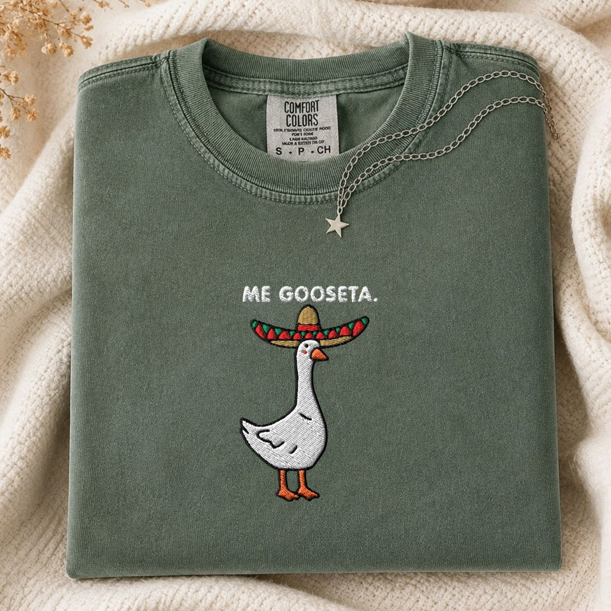 A folded sage green Comfort Colors t-shirt featuring a white goose embroidered with a colorful sombrero and the text 'ME GOOSETA.' on the front, styled on a cream knit blanket with a silver star necklace.