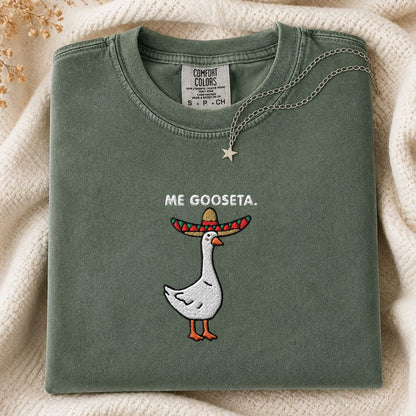 A folded sage green Comfort Colors t-shirt featuring a white goose embroidered with a colorful sombrero and the text 'ME GOOSETA.' on the front, styled on a cream knit blanket with a silver star necklace.