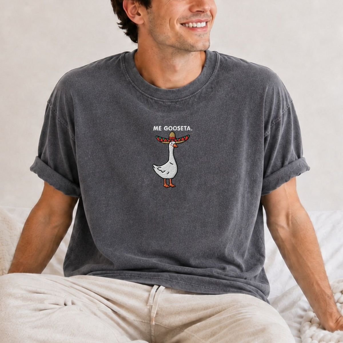 A man wearing a dark gray washed t-shirt with a cartoon goose wearing a sombrero graphic and the text 'ME GOOSETA.' sitting on a bed.