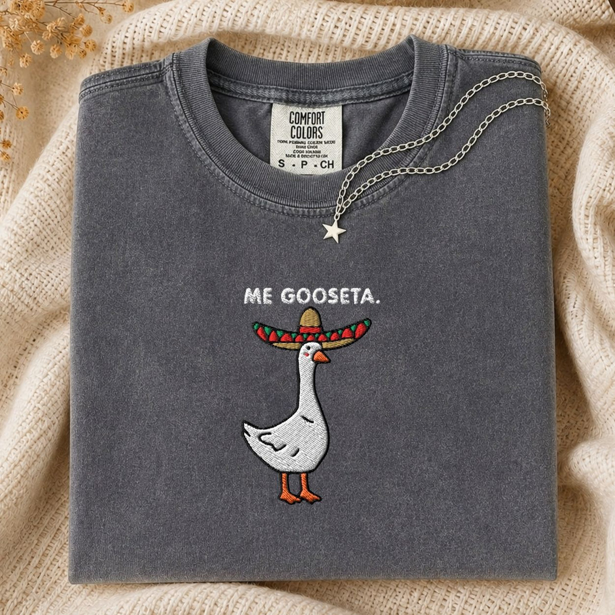 A folded dark gray Comfort Colors t-shirt with a graphic of a goose wearing a colorful sombrero and the text 'ME GOOSETA.' on the front, styled on a beige knit blanket with a silver necklace.