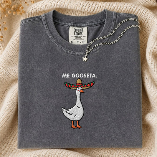 A folded dark gray Comfort Colors t-shirt with a graphic of a goose wearing a colorful sombrero and the text 'ME GOOSETA.' on the front, styled on a beige knit blanket with a silver necklace.
