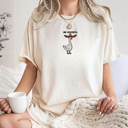 A woman wearing an oversized cream-colored t-shirt with a cartoon goose graphic wearing a sombrero and the text is 'Me Gooseta'. She is sitting in a cozy indoor setting with a chunky knit blanket and holding a white mug.