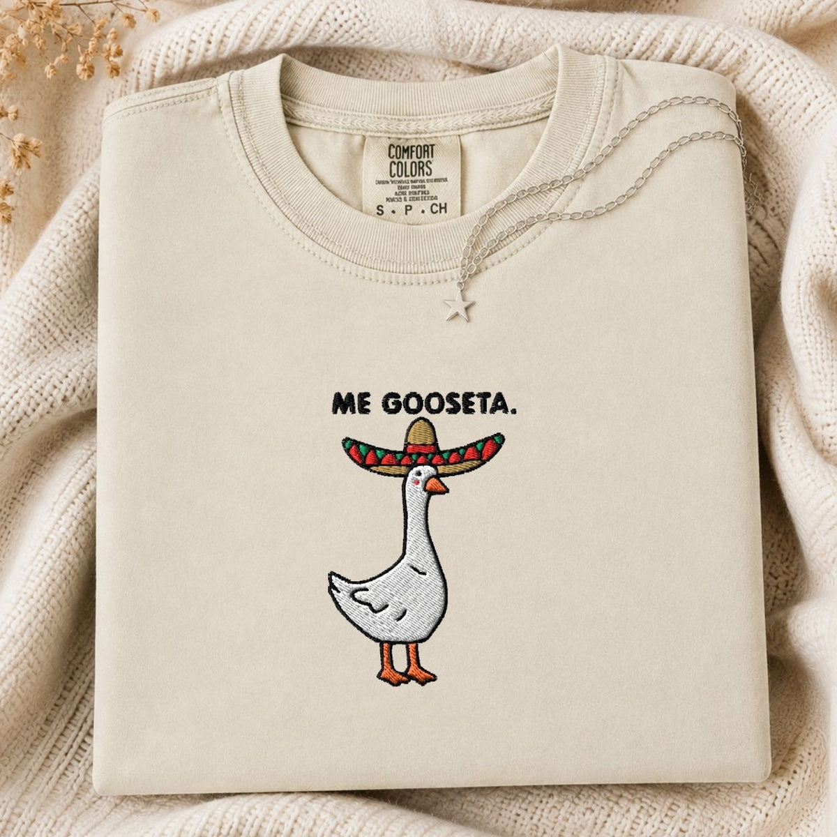 A folded cream-colored tshirt with a graphic of a white goose wearing a red and green sombrero, with the text 'ME GOOSETA.' above it.