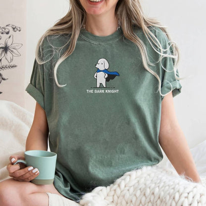 A woman wearing a sage green graphic t-shirt with a cartoon poodle superhero and the text 'THE BARK KNIGHT'. She is holding a mint green mug and sitting on a white knit blanket.