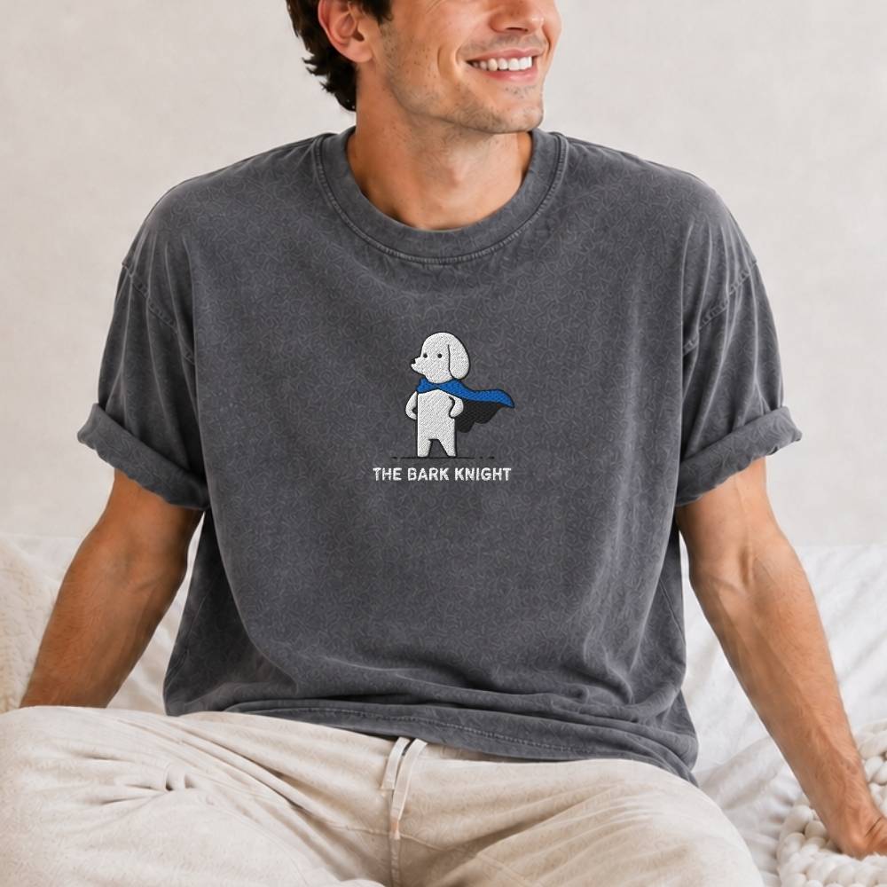 A man wearing a dark gray t-shirt with a cartoon dog superhero graphic and the text 'THE BARK KNIGHT' on the front, paired with beige pants, sitting on a bed.