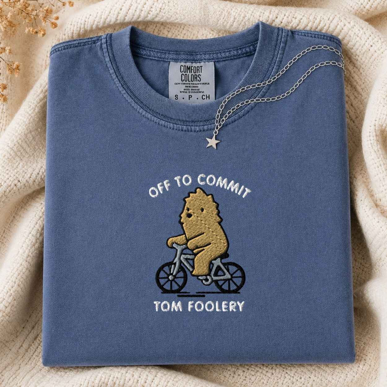 A folded slate blue t-shirt with a graphic of a bigfoot riding a bicycle and the text 'OFF TO COMMIT TOM FOOLERY'. A silver necklace with a star pendant is draped over the collar.