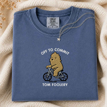 A folded slate blue t-shirt with a graphic of a bigfoot riding a bicycle and the text 'OFF TO COMMIT TOM FOOLERY'. A silver necklace with a star pendant is draped over the collar.
