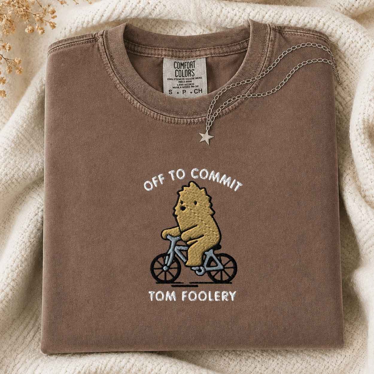 A folded brown crewneck tshirt with a graphic of a bigfoot riding a bicycle and the text 'OFF TO COMMIT TOM FOOLERY', styled on a cream knit blanket with a silver necklace.