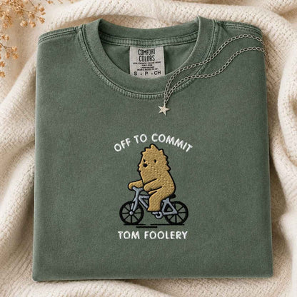 A folded sage green t-shirt with a graphic of a cartoon bigfoot riding a bicycle and the text 'OFF TO COMMIT TOM FOOLERY bigfoot'. A silver necklace with a star pendant is draped over the collar.