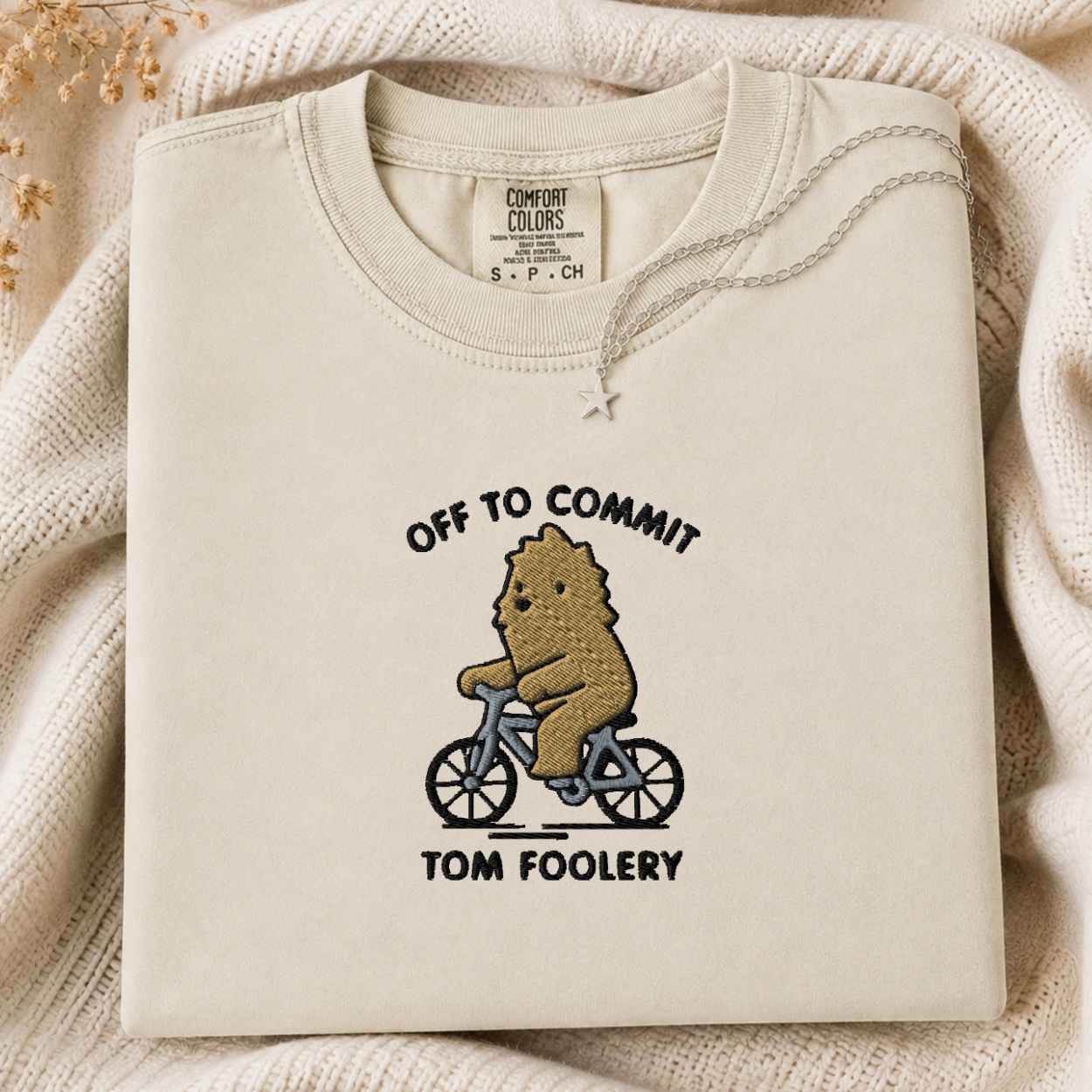 A folded cream-colored crewneck tshirt with a graphic of a brown bear riding a blue bicycle and the text 'OFF TO COMMIT TOM FOOLERY'. The tshirt is displayed on a cozy, textured cream knit blanket with dried flowers in the corner.
