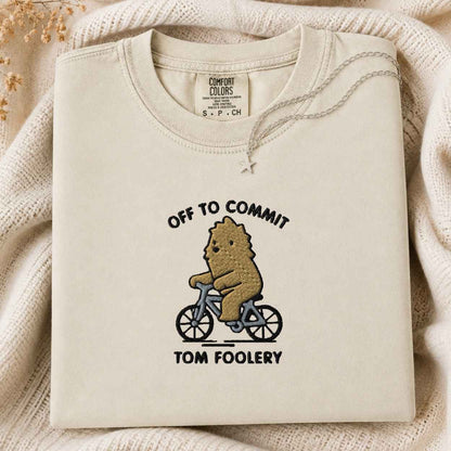 A folded cream-colored crewneck tshirt with a graphic of a brown bear riding a blue bicycle and the text 'OFF TO COMMIT TOM FOOLERY'. The tshirt is displayed on a cozy, textured cream knit blanket with dried flowers in the corner.