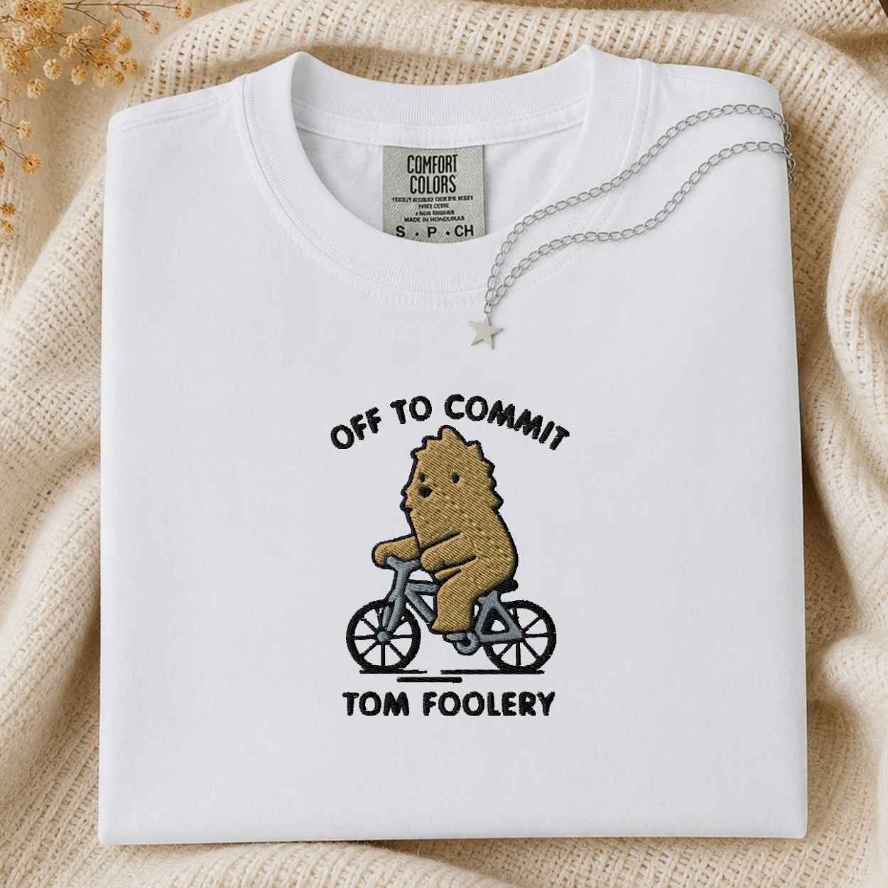 A folded white Comfort Colors t-shirt with a graphic of a bigfoot riding a bicycle and the text 'OFF TO COMMIT TOM FOOLERY'. A silver necklace with a star pendant is draped over the collar.