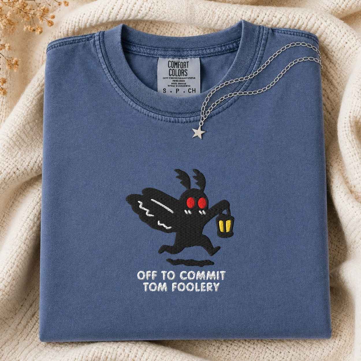 A folded slate blue t-shirt with a graphic of a black moth cartoon character with red eyes holding a lantern, with the text 'OFF TO COMMIT TOM FOOLERY' printed below. A silver necklace with a star pendant is draped over the collar.