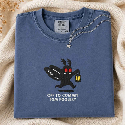 A folded slate blue t-shirt with a graphic of a black moth cartoon character with red eyes holding a lantern, with the text 'OFF TO COMMIT TOM FOOLERY' printed below. A silver necklace with a star pendant is draped over the collar.