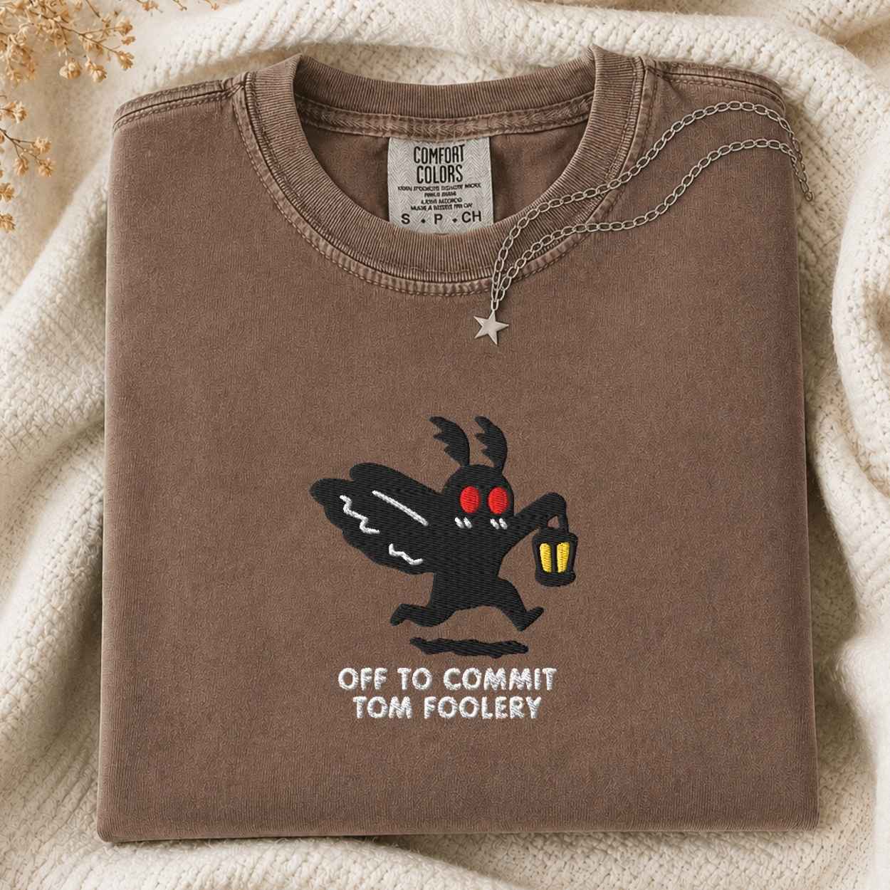 A folded brown t-shirt with a graphic of the moth character Off to Commit Tom Foolery, styled on a cream knit blanket with a silver necklace.