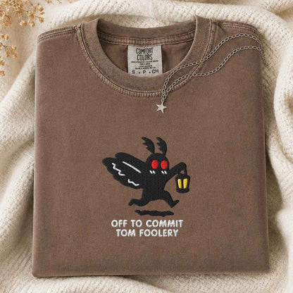 A folded brown t-shirt with a graphic of the moth character Off to Commit Tom Foolery, styled on a cream knit blanket with a silver necklace.