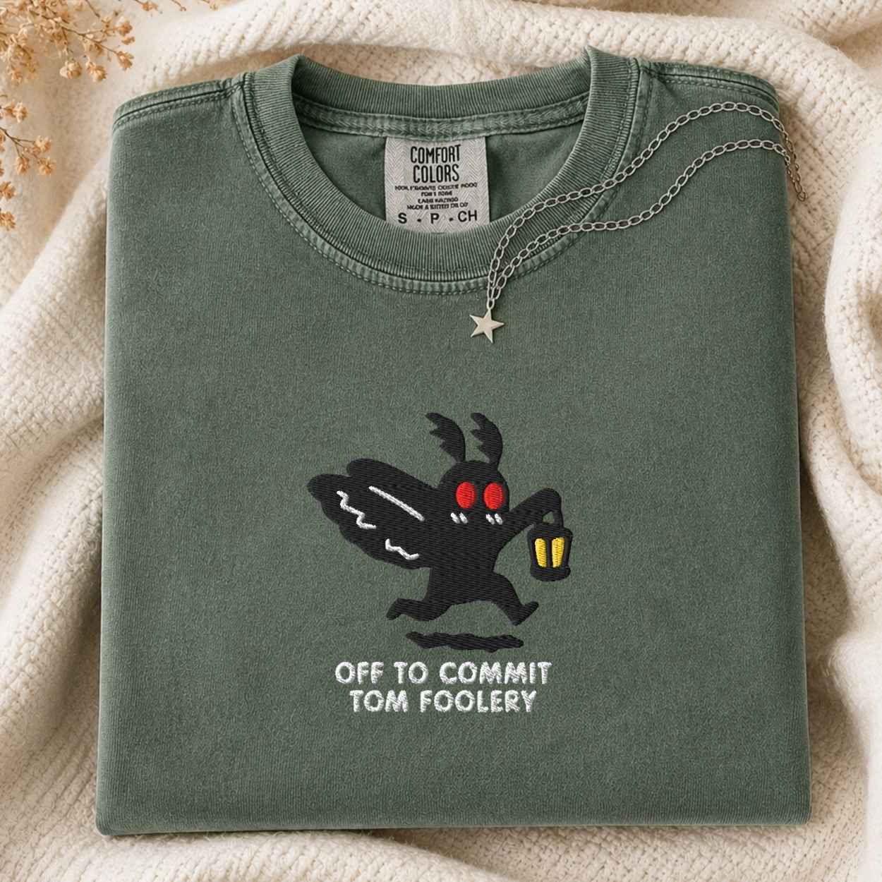A folded sage green Comfort Colors t-shirt featuring a black moth cartoon character with red eyes and the text 'OFF TO COMMIT TOM FOOLERY' on the front, styled on a cream knit blanket with a silver necklace.