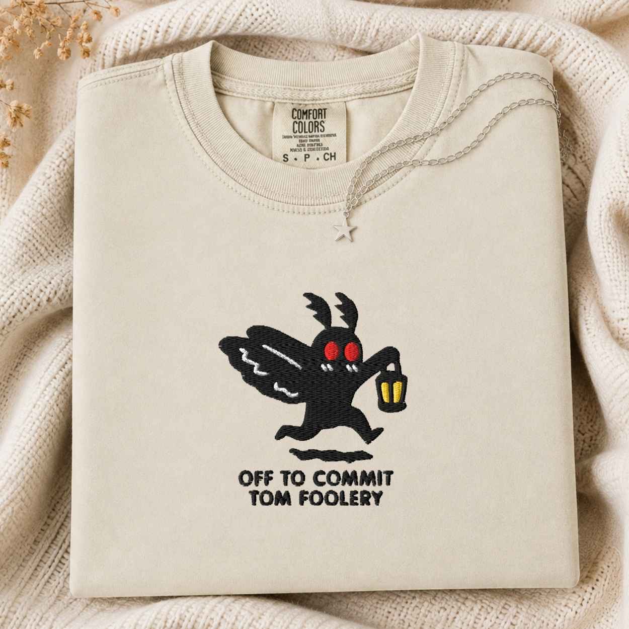 A folded cream-colored sweatshirt with a graphic of the Tom Foolery character and the text 'OFF TO COMMIT TOM FOOLERY' printed on the front, styled on a cozy cream knit blanket.