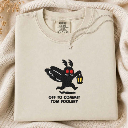 A folded cream-colored sweatshirt with a graphic of the Tom Foolery character and the text 'OFF TO COMMIT TOM FOOLERY' printed on the front, styled on a cozy cream knit blanket.