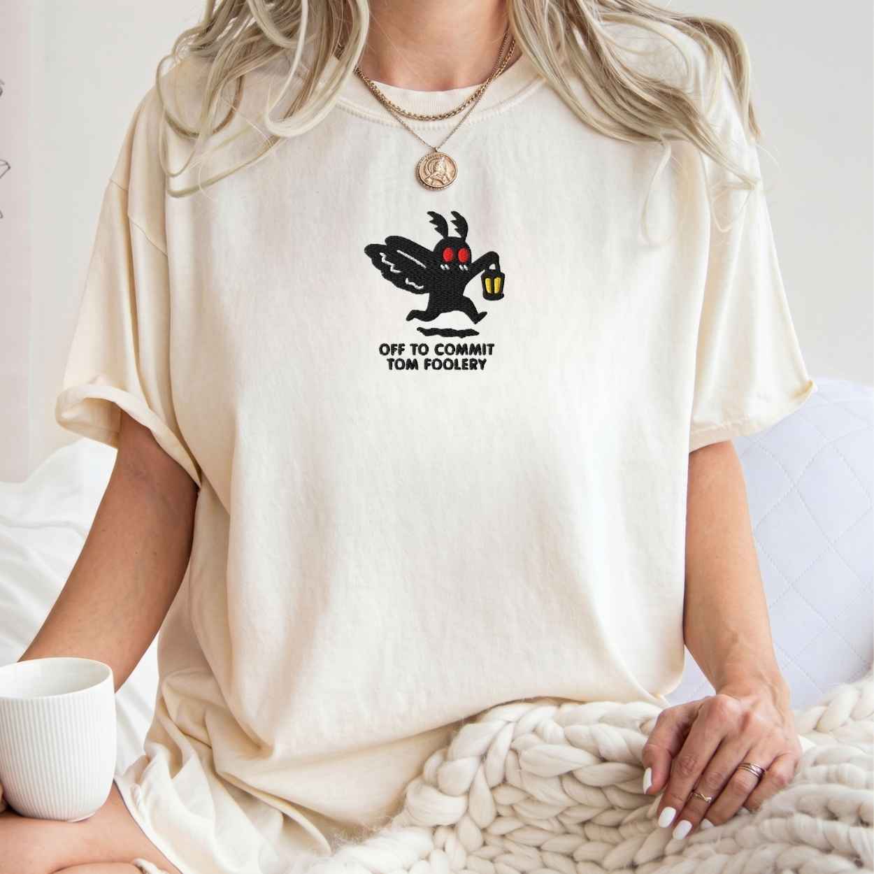 A woman wearing an oversized cream-colored t-shirt with a black cartoon moth graphic and the text 'OFF TO COMMIT TOM FOOLERY' printed on the chest. She is sitting on a white couch with a chunky knit blanket and a white mug in the foreground.