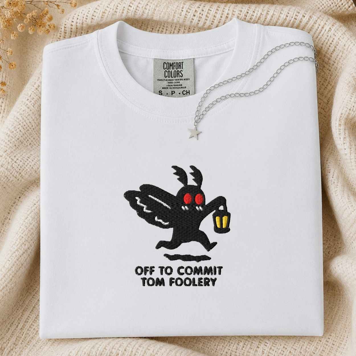 A folded white Comfort Colors t-shirt featuring a black moth cartoon character with red eyes holding a yellow lantern, with the text 'OFF TO COMMIT TOM FOOLERY' printed below. The shirt is styled on a beige knit blanket with a silver necklace draped over the collar.