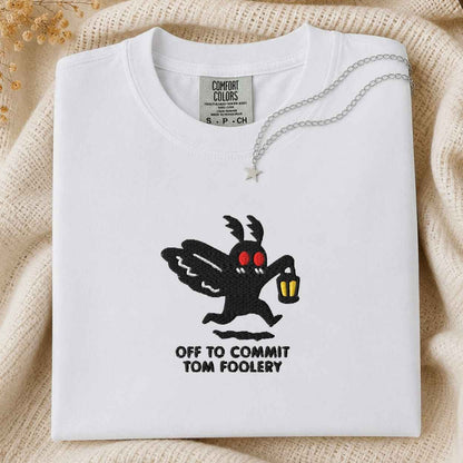 A folded white Comfort Colors t-shirt featuring a black moth cartoon character with red eyes holding a yellow lantern, with the text 'OFF TO COMMIT TOM FOOLERY' printed below. The shirt is styled on a beige knit blanket with a silver necklace draped over the collar.