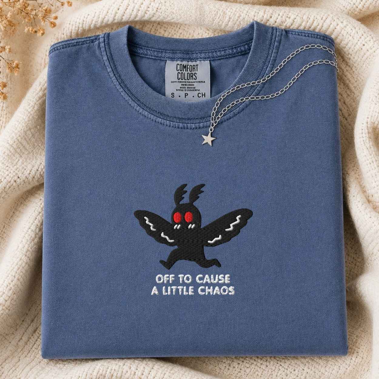 A folded slate blue Comfort Colors t-shirt with a black embroidered moth graphic, red eyes, and the text 'OFF TO CAUSE A LITTLE CHAOS' in white. A silver necklace with a star pendant is draped over the collar.