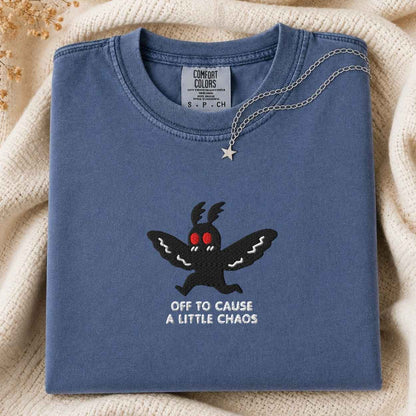 A folded slate blue Comfort Colors t-shirt with a black embroidered moth graphic, red eyes, and the text 'OFF TO CAUSE A LITTLE CHAOS' in white. A silver necklace with a star pendant is draped over the collar.