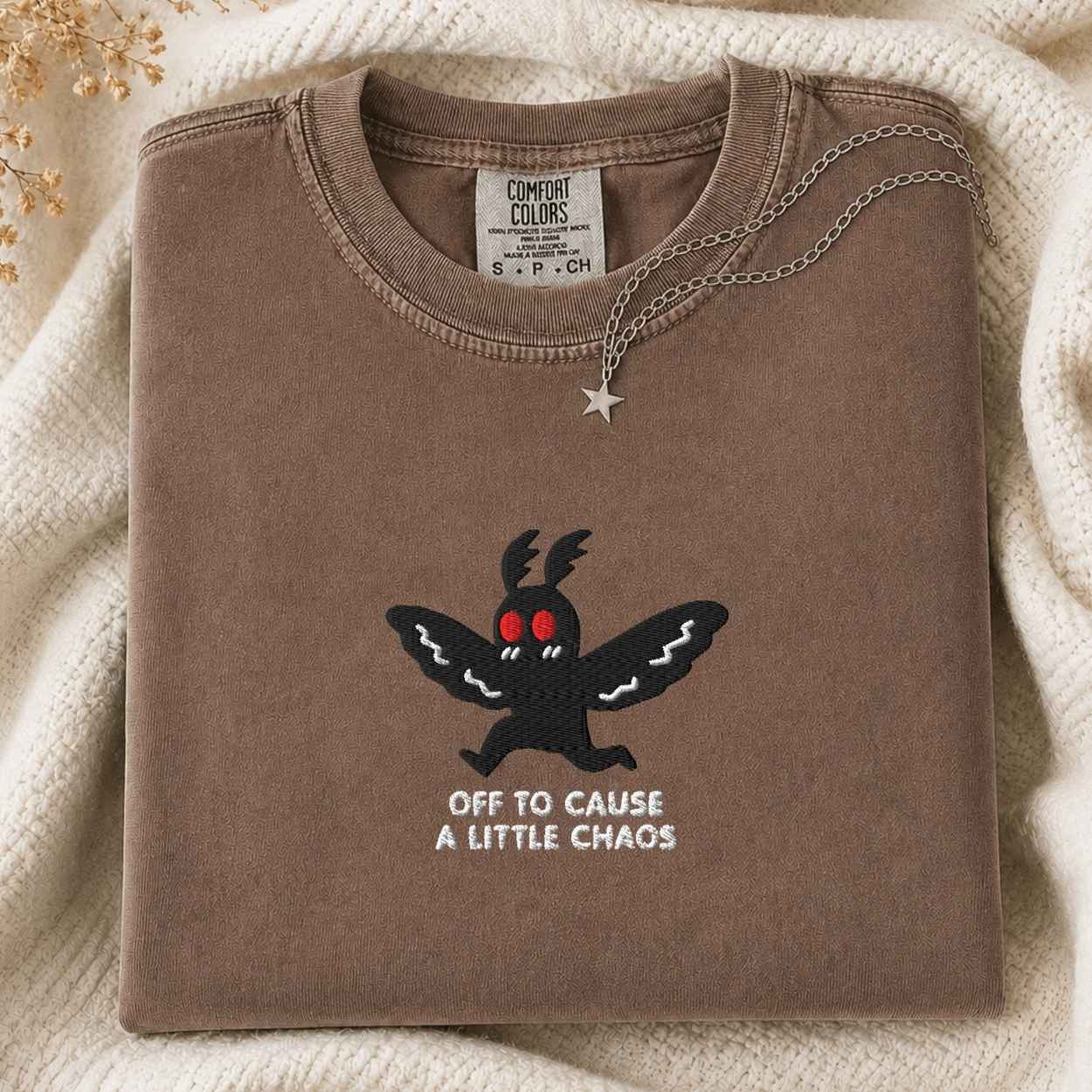 A folded brown t-shirt with a black cartoon moth graphic and the text 'OFF TO CAUSE A LITTLE CHAOS' on the front. The shirt is styled on a cream-colored knit blanket with a silver star necklace draped over the collar.