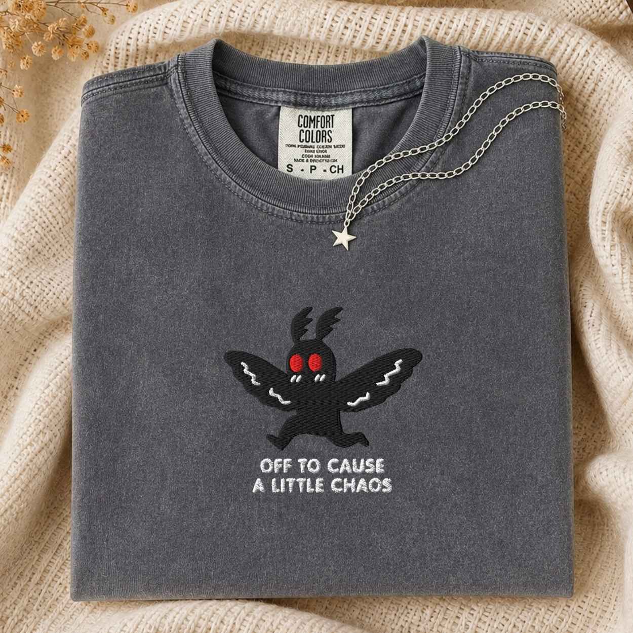 A folded dark gray Comfort Colors t-shirt with a black moth graphic and the text 'OFF TO CAUSE A LITTLE CHAOS' in white, styled on a beige knit blanket with a silver star necklace draped over it.
