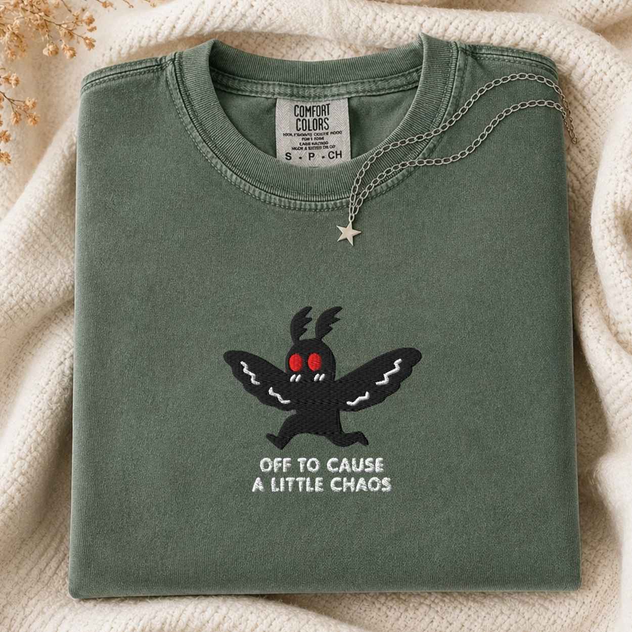 A folded sage green Comfort Colors t-shirt featuring a black embroidered moth with red eyes and the text 'OFF TO CAUSE A LITTLE CHAOS' in white. The shirt is styled on a cream knit blanket with a silver star necklace draped over the collar.