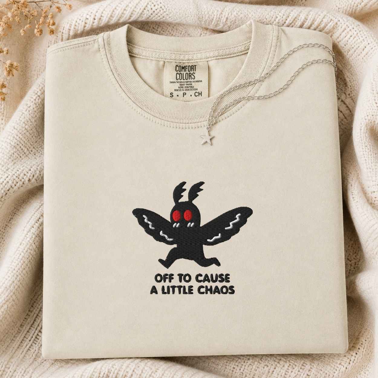 A folded cream-colored sweatshirt with a black cartoon moth graphic and the text 'OFF TO CAUSE A LITTLE CHAOS' on the front, displayed on a cozy cream knit blanket.