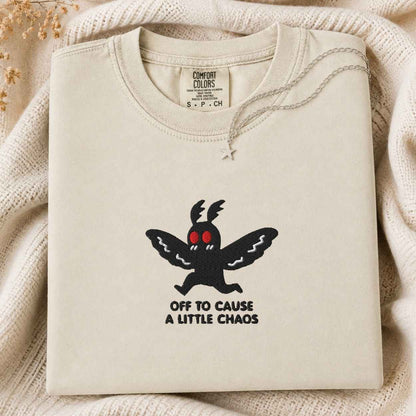 A folded cream-colored sweatshirt with a black cartoon moth graphic and the text 'OFF TO CAUSE A LITTLE CHAOS' on the front, displayed on a cozy cream knit blanket.