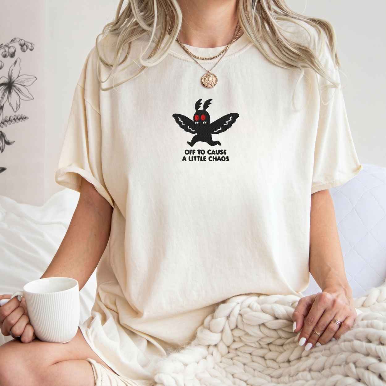A woman wearing a cream-colored oversized t-shirt with a black cartoon moth graphic and the text 'OFF TO CAUSE A LITTLE CHAOS'. She is sitting in a cozy room setting, holding a white mug and wrapped in a chunky knit blanket.