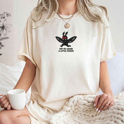 A woman wearing a cream-colored oversized t-shirt with a black cartoon moth graphic and the text 'OFF TO CAUSE A LITTLE CHAOS'. She is sitting in a cozy room setting, holding a white mug and wrapped in a chunky knit blanket.