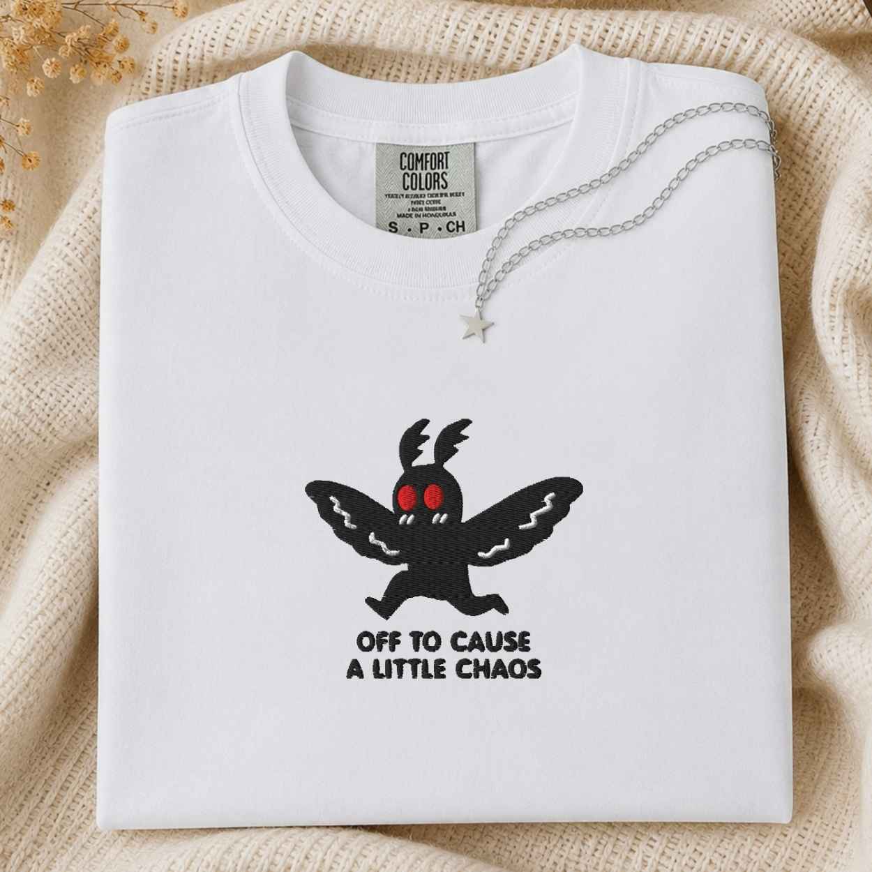 A folded white t-shirt with a black graphic of a winged creature with red eyes and the text 'OFF TO CAUSE A LITTLE CHAOS'. A silver necklace with a star pendant is draped over the collar.