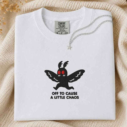 A folded white t-shirt with a black graphic of a winged creature with red eyes and the text 'OFF TO CAUSE A LITTLE CHAOS'. A silver necklace with a star pendant is draped over the collar.