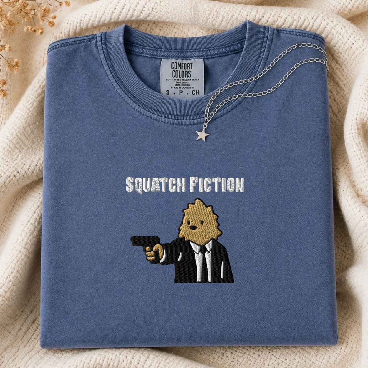 A folded slate blue t-shirt with a graphic of a squatch in a suit holding a gun and the text 'SQUATCH FICTION' embroidered on the front. The shirt is styled on a cream-colored knit blanket with a silver necklace draped over the collar.
