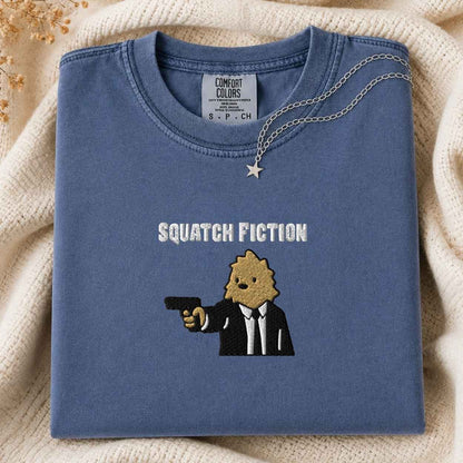 A folded slate blue t-shirt with a graphic of a squatch in a suit holding a gun and the text 'SQUATCH FICTION' embroidered on the front. The shirt is styled on a cream-colored knit blanket with a silver necklace draped over the collar.
