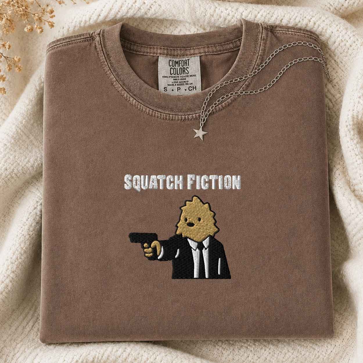 A folded brown tshirt with a graphic print reading 'SQUATCH FICTION' and showing a cartoon bigfoot in a black suit holding a gun, styled on a cream knit blanket with a small star necklace.