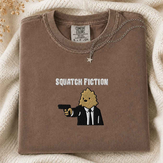 A folded brown tshirt with a graphic print reading 'SQUATCH FICTION' and showing a cartoon bigfoot in a black suit holding a gun, styled on a cream knit blanket with a small star necklace.