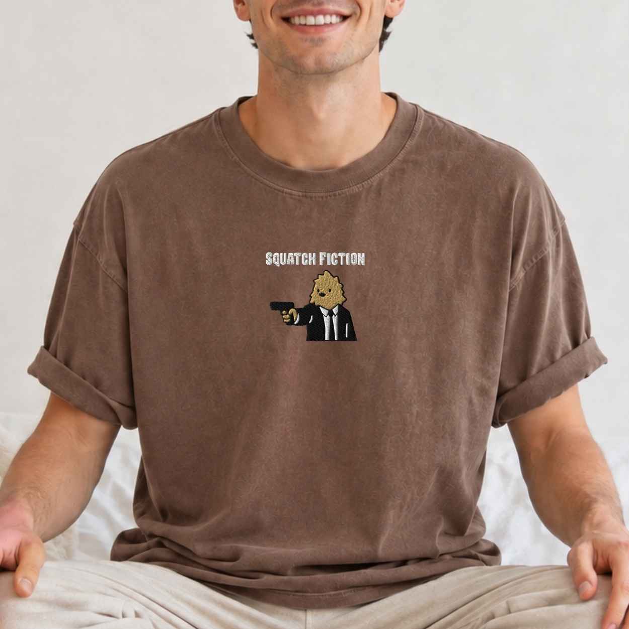 A man wearing a brown crewneck t-shirt with a graphic of a cartoon bigfoot in a suit holding a gun and the text 'SQUATCH FICTION'.