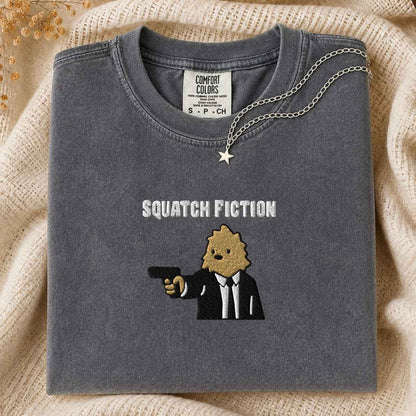 A folded dark gray Comfort Colors t-shirt with a 'Squatch Fiction' graphic of a bigfoot in a suit holding a gun, styled on a beige knit blanket with a silver star necklace.